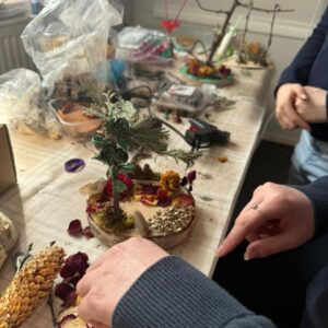 Naturmaterialien-Workshop: Hände gestalten ein Miniatur-Bäumchen auf einer Holzscheibe mit getrockneten Blüten.