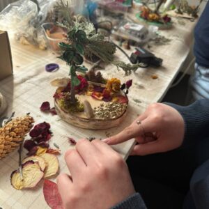 Kunsttherapie-Prozess: Hände dekorieren ein kreatives Miniatur-Gesteck mit getrockneten Rosenblättern, Moos und Zapfen auf einem Holztisch bei Farbe und Klang Leipzig.